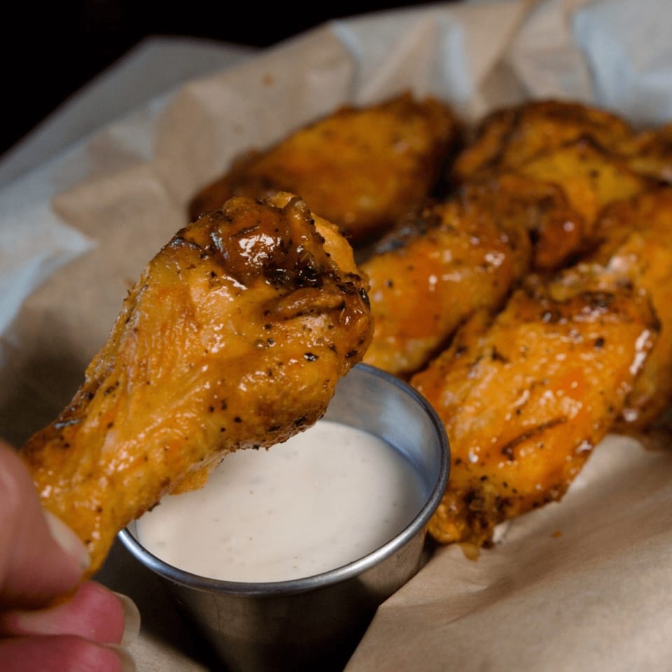A hand dips a golden-brown chicken wing into a small metal cup of white sauce at Pizza On Tap.