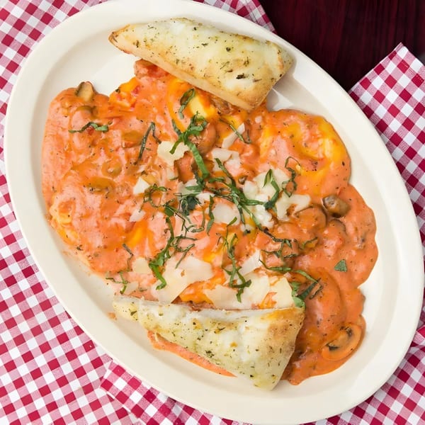 Creamy tomato pasta with mushrooms, basil, and garlic bread on a white plate and red checkered tablecloth.