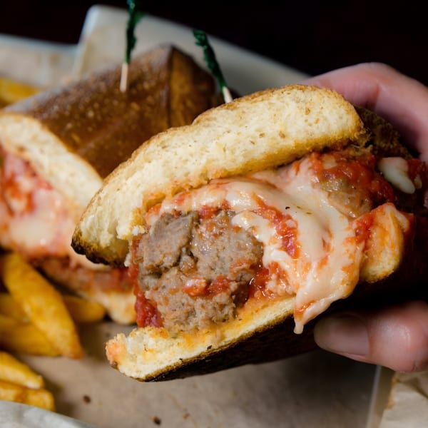 Hand holding a meatball sub sandwich with melted cheese and tomato sauce, with fries in the background.