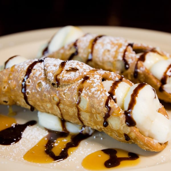 Two cannolis filled with cream, drizzled with chocolate and caramel, dusted with powdered sugar.