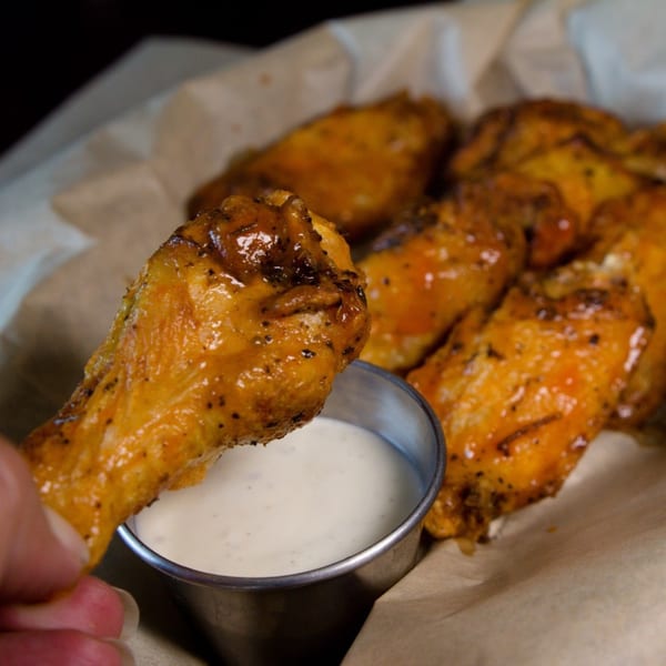 A hand dips a golden-brown chicken wing into a small metal cup of white dipping sauce.