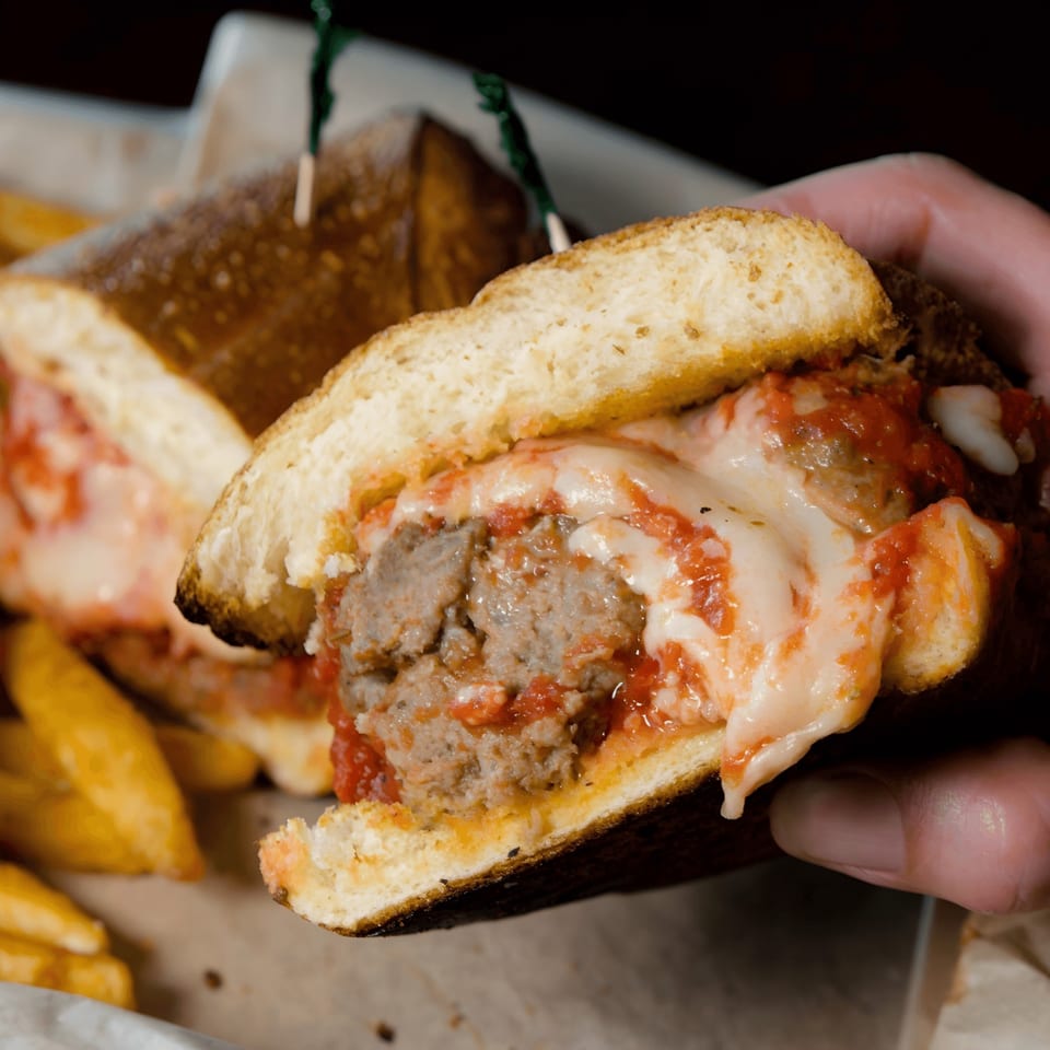 Hand holding a meatball sub sandwich with melted cheese and marinara sauce, with fries in the background.